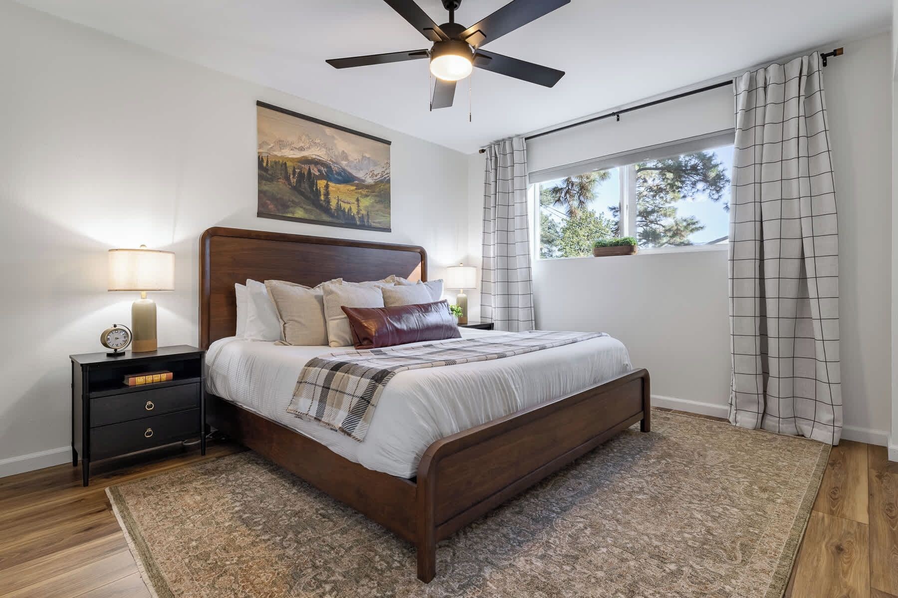 A bedroom with a bed, a nightstand, a ceiling fan, a window with curtains, and a painting on the wall.