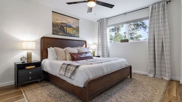 A bedroom with a bed, a nightstand, a ceiling fan, a window with curtains, and a painting on the wall.