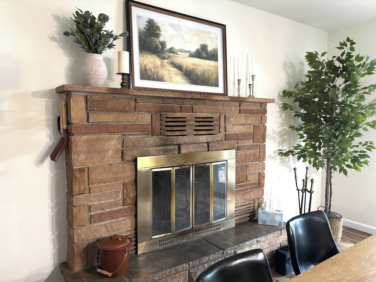 A fireplace with a stone mantle, a framed landscape painting above it, and a potted plant to the right.