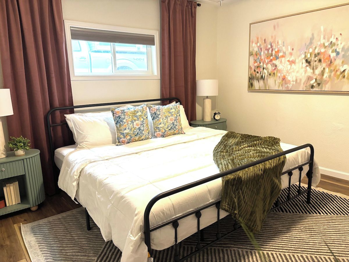 A bedroom with a bed, a bedside table, a lamp, a window with curtains, and a painting on the wall.