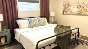 A bedroom with a bed, a bedside table, a lamp, a window with curtains, and a painting on the wall.