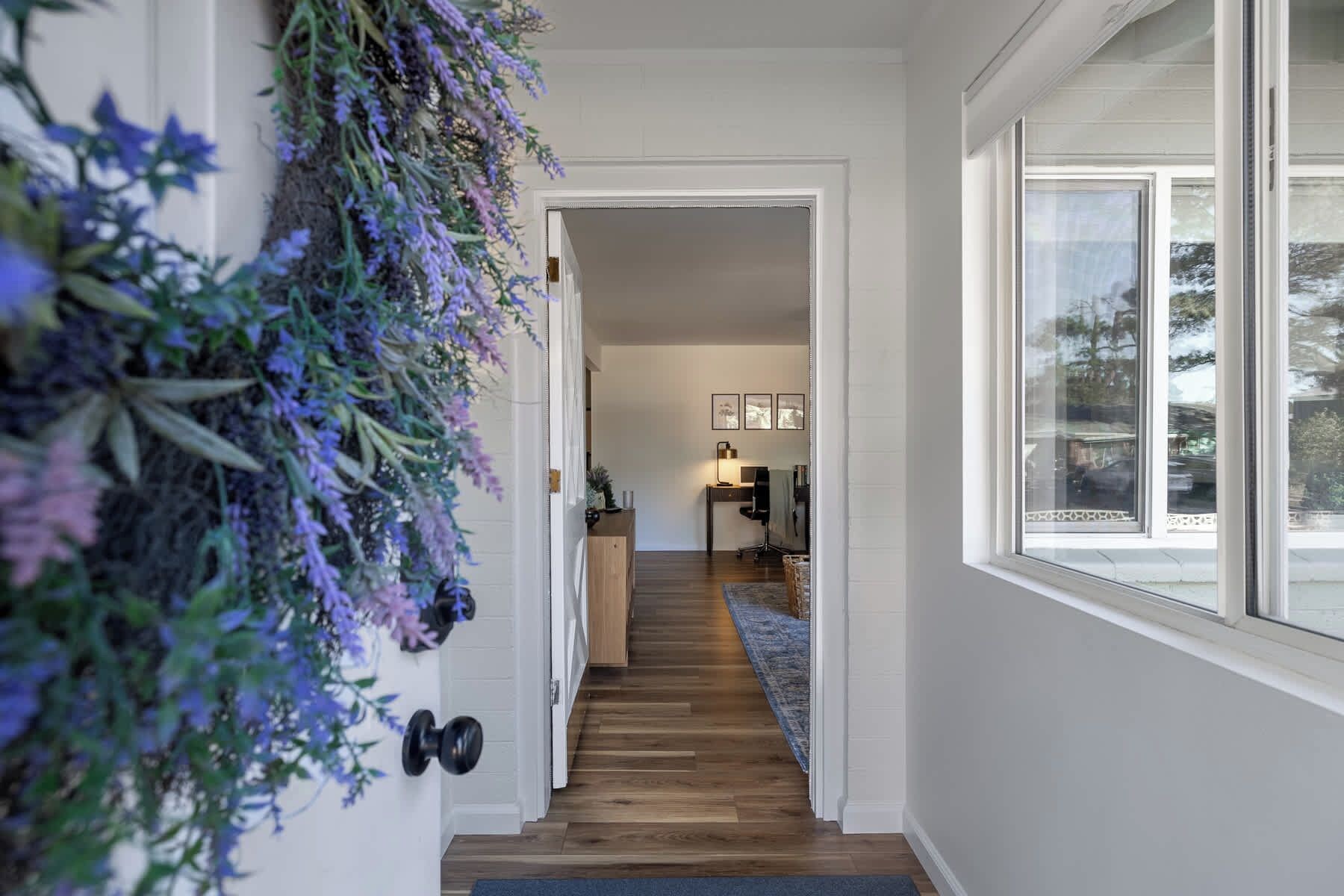 Un couloir avec un escalier en bois menant à une pièce où se trouvent un bureau et une chaise, une fenêtre donnant sur des arbres et une plante fixée au mur, ornée de fleurs violettes.
