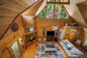 A cozy cabin interior with a stone fireplace, wooden walls, and a large window offering a view of trees. - Lake Lure Lakefront Home with Private Par 3 (Lake Lure)