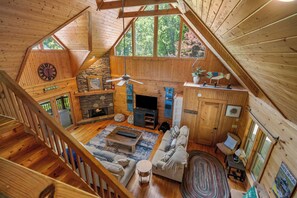 A spacious loft-style room with a stone fireplace, wooden floors, a ceiling fan, and large windows offering a view of the outdoors. - Lake Lure Lakefront Home with Private Par 3 (Lake Lure)