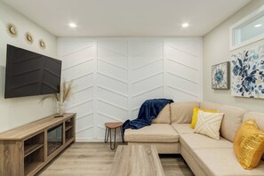Uma sala de estar moderna com sofá seccional, televisão de tela plana, mesa de centro de madeira e um painel decorativo na parede.