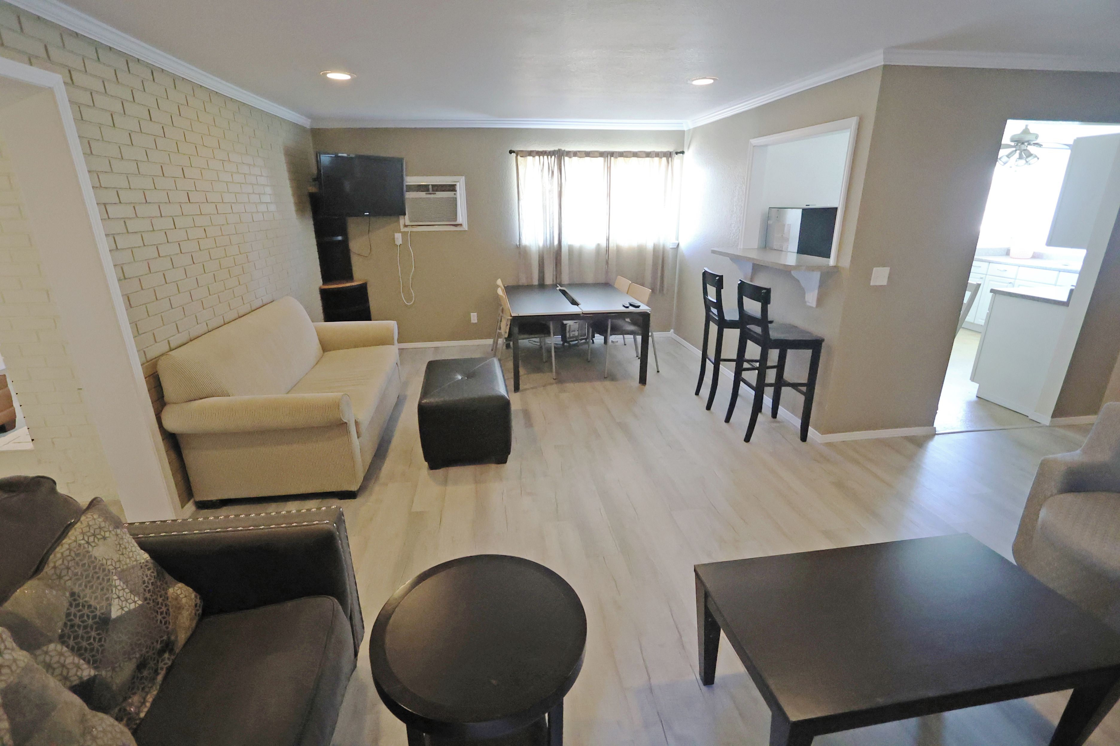 A living room with a sofa, a coffee table, and a TV. There is a dining area with a table and chairs. The room has a brick wall and a window with curtains.