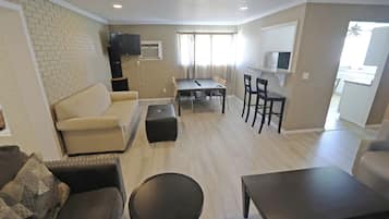 A living room with a sofa, a coffee table, and a TV. There is a dining area with a table and chairs. The room has a brick wall and a window with curtains.