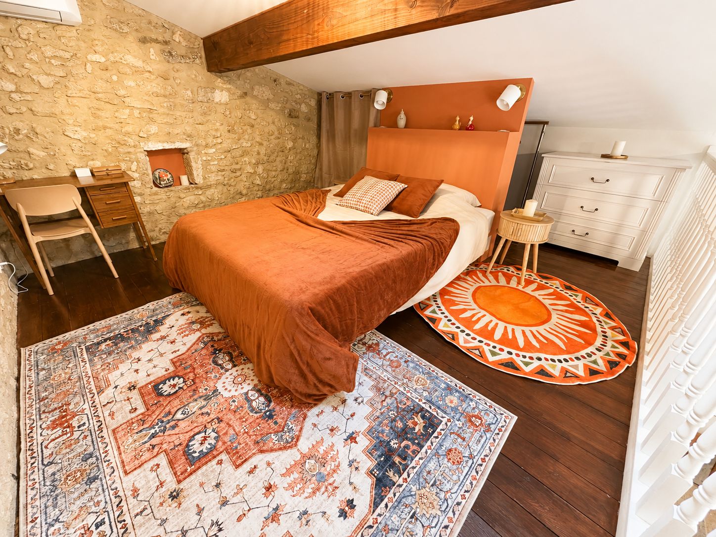 Une chambre avec un mur en pierre, un cadre de lit en bois, un tapis à motifs et une commode blanche.