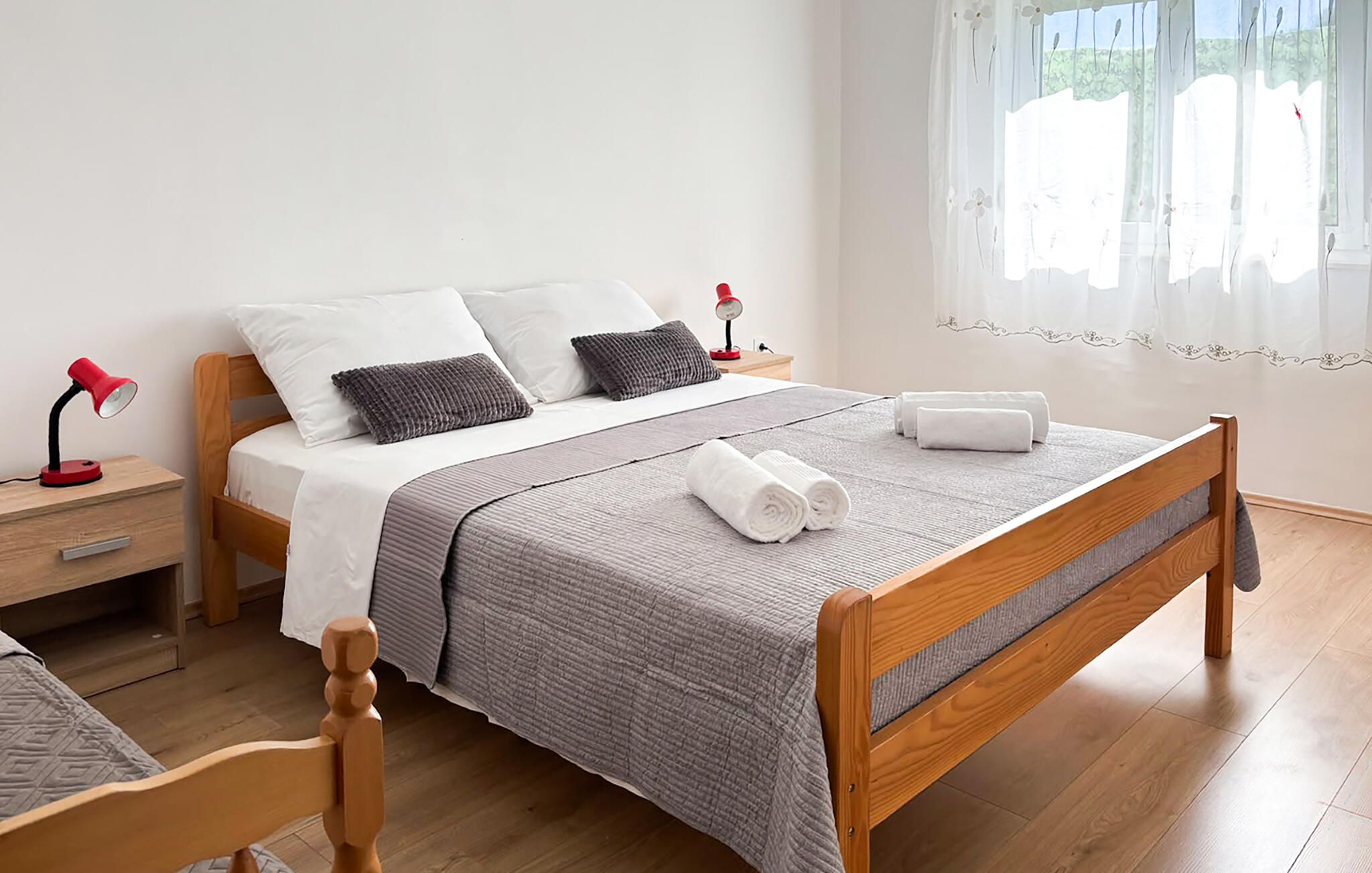 A neatly made bed with a grey blanket and two pillows, a wooden nightstand with a red lamp, and a window with sheer curtains.