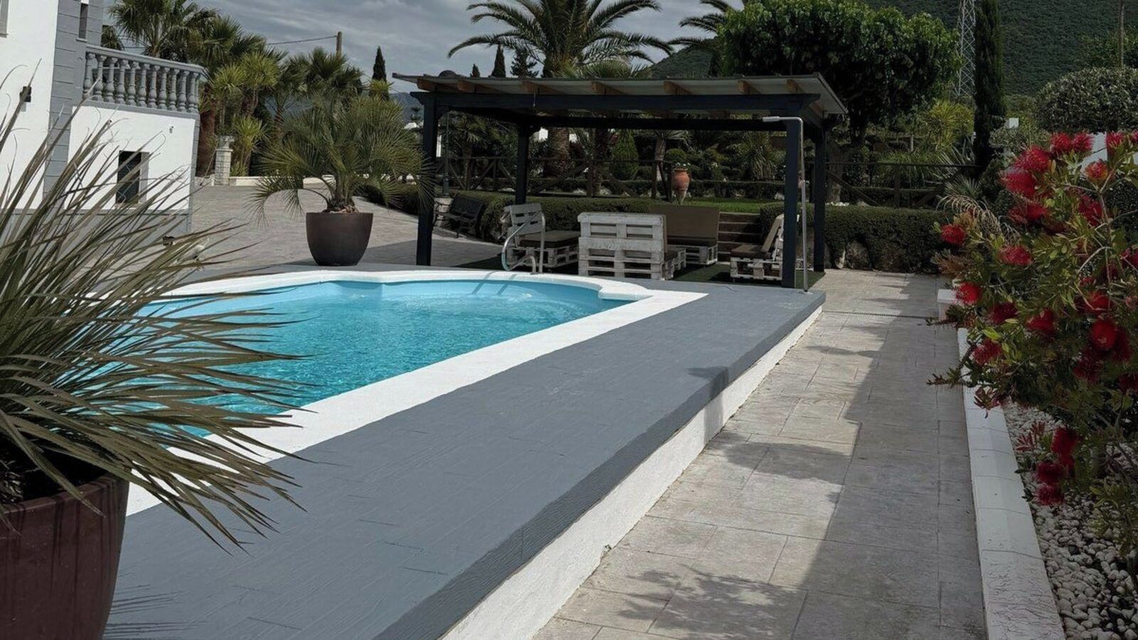 A rectangular swimming pool with a white edge, surrounded by a paved deck. There are outdoor seating arrangements and a pergola structure in the background.