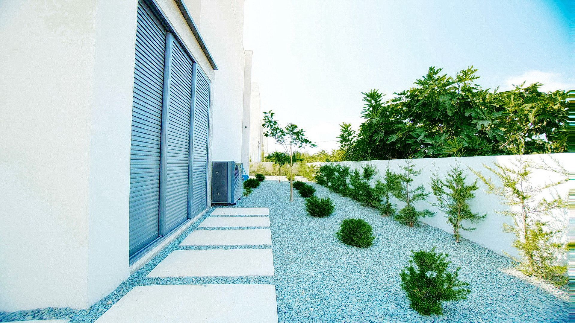 Een modern gebouw met een witte gevel, een blauwe deur en een grindpad dat leidt naar een kleine tuin met jonge bomen.