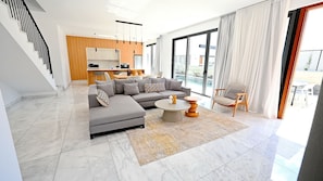 A modern living room with a sectional sofa, a coffee table, and a chair. There is a kitchen area in the background with a bar and a dining table.
