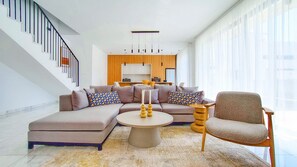 A modern living room with a sectional sofa, a round coffee table, and a chair. There is a staircase with a railing on the left, and a kitchen area in the background. - Villa Allegra Elite - Luxury Villa In Kato Paphos (Paphos)
