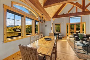 A spacious dining area with a long wooden table, multiple chairs, and large windows offering views of a forested landscape. - Willow Lodge, Pet friendly, deck, garage. (Crested Butte)
