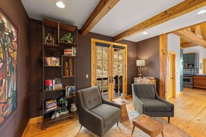A spacious living room with a wooden floor, two grey armchairs, a bookshelf, and a dining area visible through a glass door. - Willow Lodge, Pet friendly, deck, garage. (Crested Butte)