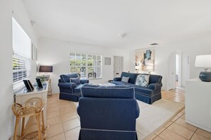 Ein geräumiges Wohnzimmer mit einem Ecksofa, einem Couchtisch und einem Fernseher. Es gibt Fenster mit Jalousien, eine Tür und eine Lampe auf einem Beistelltisch.