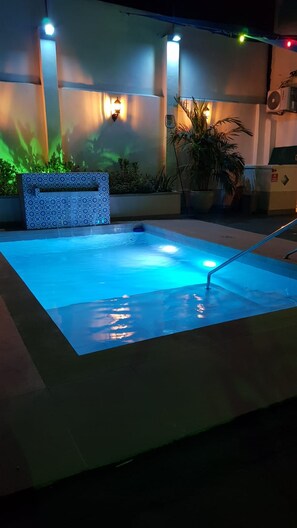Una piscina con una fuente iluminada en azul, rodeada por un solado de baldosas y plantas en macetas.