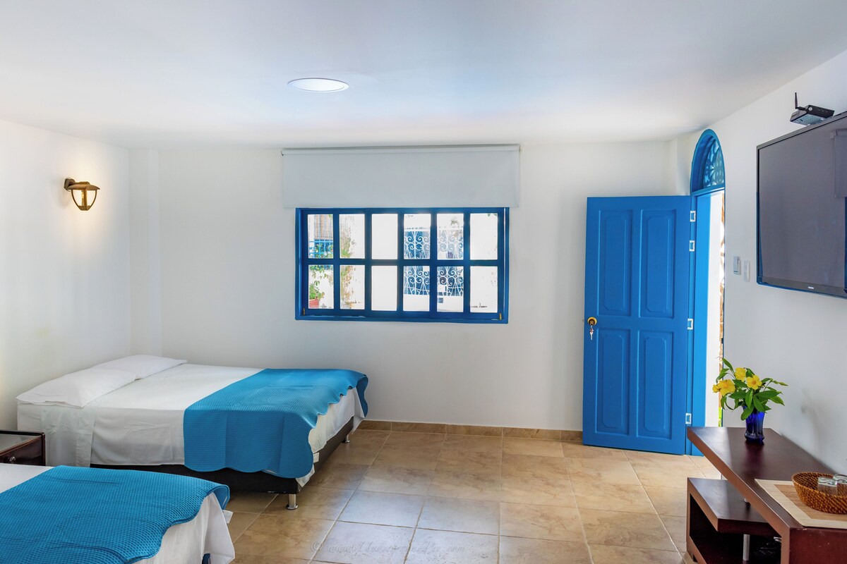 Habitació d'hotel amb dos llits, una finestra amb persianes blaves, una porta blava i un televisor de pantalla plana penjat a la paret.