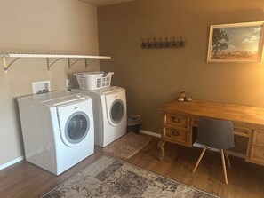 Een wasserette met twee witte Bosch wasmachines, een houten bureau met een stoel, een wandkapstok en een lijst met een landschapsafbeelding.
