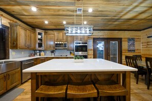 A kitchen with wooden cabinets, a central island with seating, stainless steel appliances, and a hanging light fixture. - A Beautiful View · New Listing! Ridgetop 5BR w/ Hot Tub, Game Room & Fire Pit (Broken Bow)