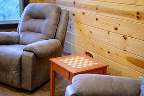 A room with a brown leather armchair, a wooden table with a chessboard, and a wooden wall. - A Beautiful View · New Listing! Ridgetop 5BR w/ Hot Tub, Game Room & Fire Pit (Broken Bow)