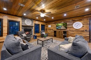 A cozy living room with a fireplace, comfortable seating, and a large TV screen. - A Beautiful View · New Listing! Ridgetop 5BR w/ Hot Tub, Game Room & Fire Pit (Broken Bow)