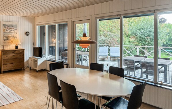 Een kamer met een ronde tafel en stoelen, grote ramen, een houten dressoir en een hangende lamp.