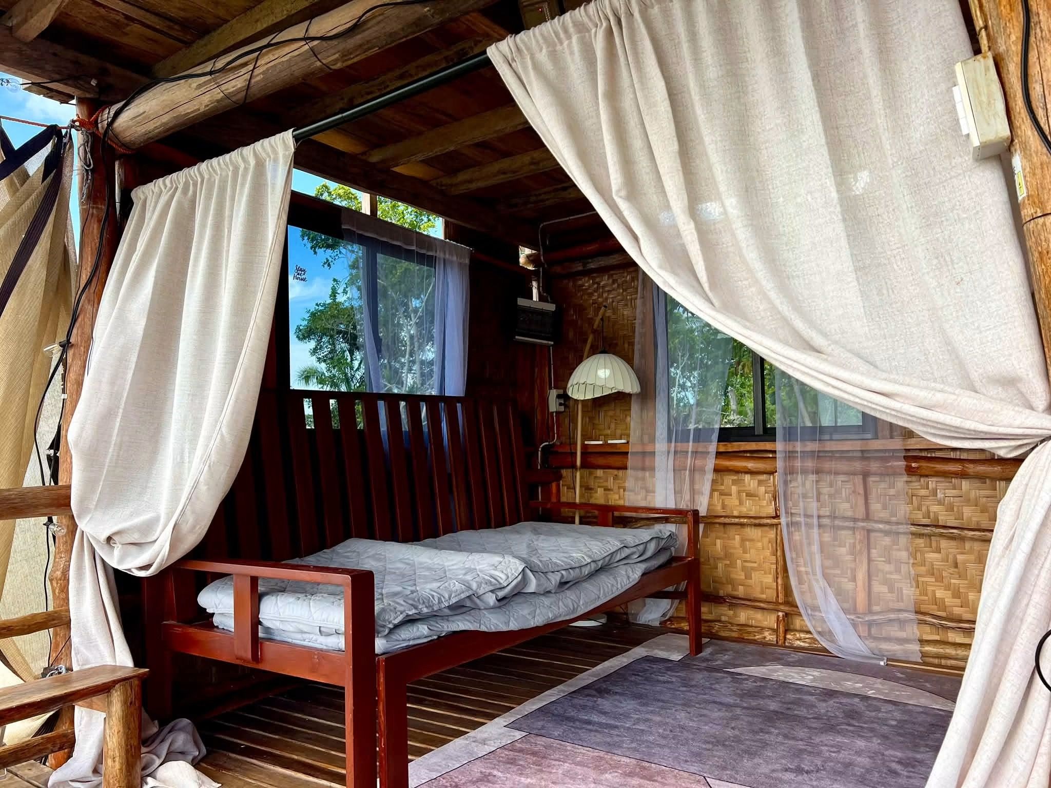 A single bed with a mosquito net, wooden walls, and a window with a view of trees.