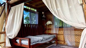 A single bed with a mosquito net, wooden walls, and a window with a view of trees.