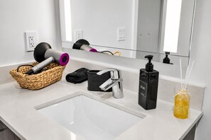 Ein Badezimmer mit einem weißen Waschbecken, einem Spiegel, einem schwarzen Seifenspender, einer gelben Flasche, einem schwarzen Handtuch und einem gewebten Korb mit einem Fön und einer schwarzen Haarbürste.