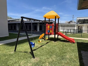 A playground with a slide and swings, surrounded by grass and a building in the background. - Azure Horizon Jaco - Beach & Ocean View Apartment (Jaco)
