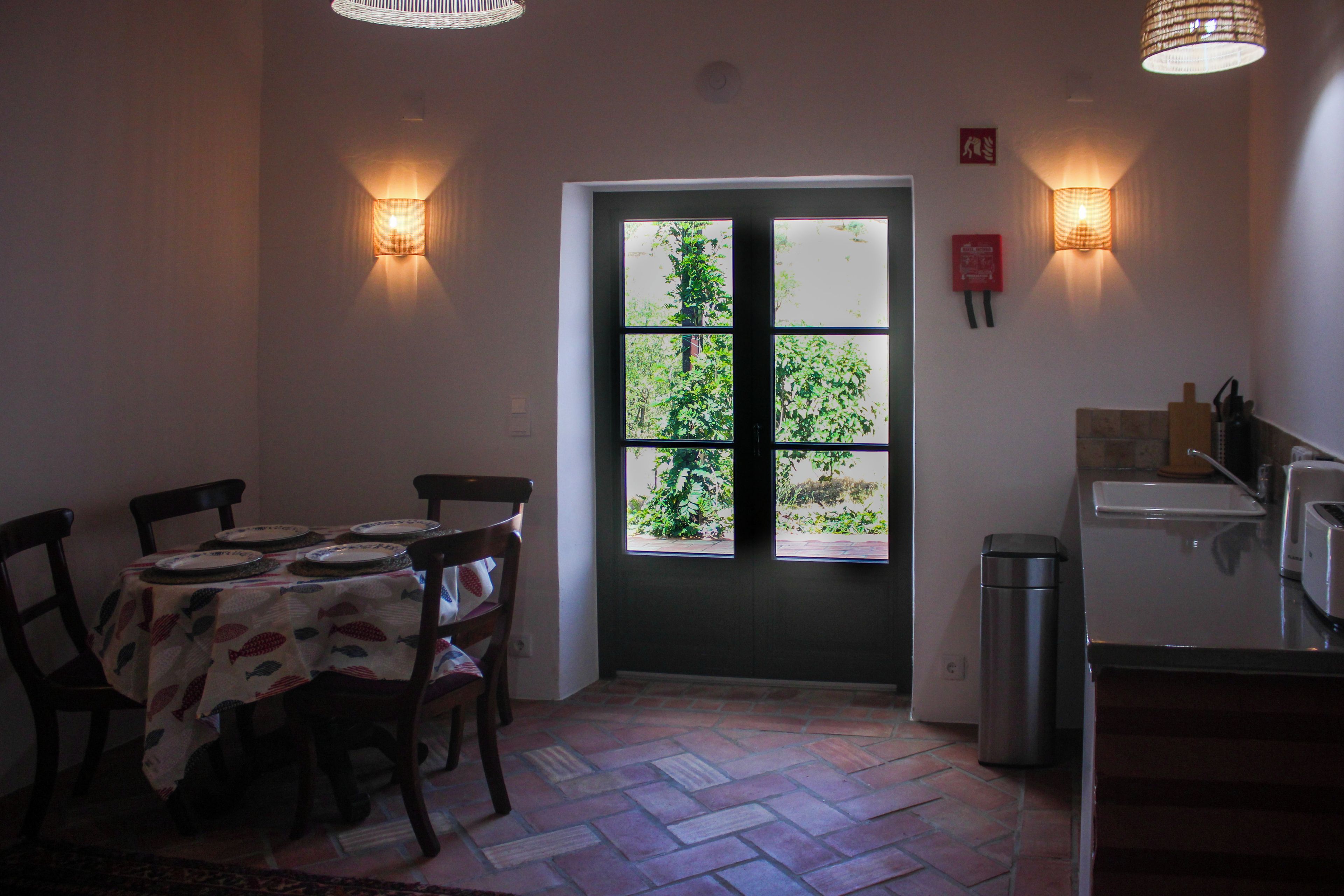 A dining area with a table set for four, a door with a view of greenery, and a kitchen area with a trash bin and a toaster.