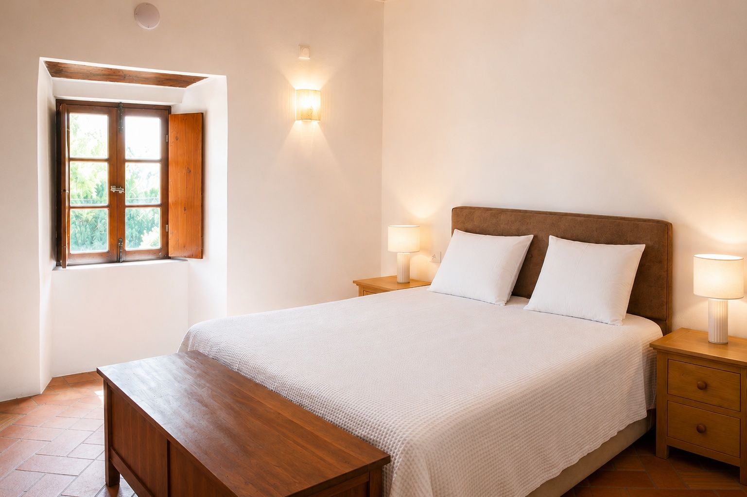 Un dormitorio con cama, dos lámparas de noche, un banco de madera, una ventana con vistas y una lámpara de pared.