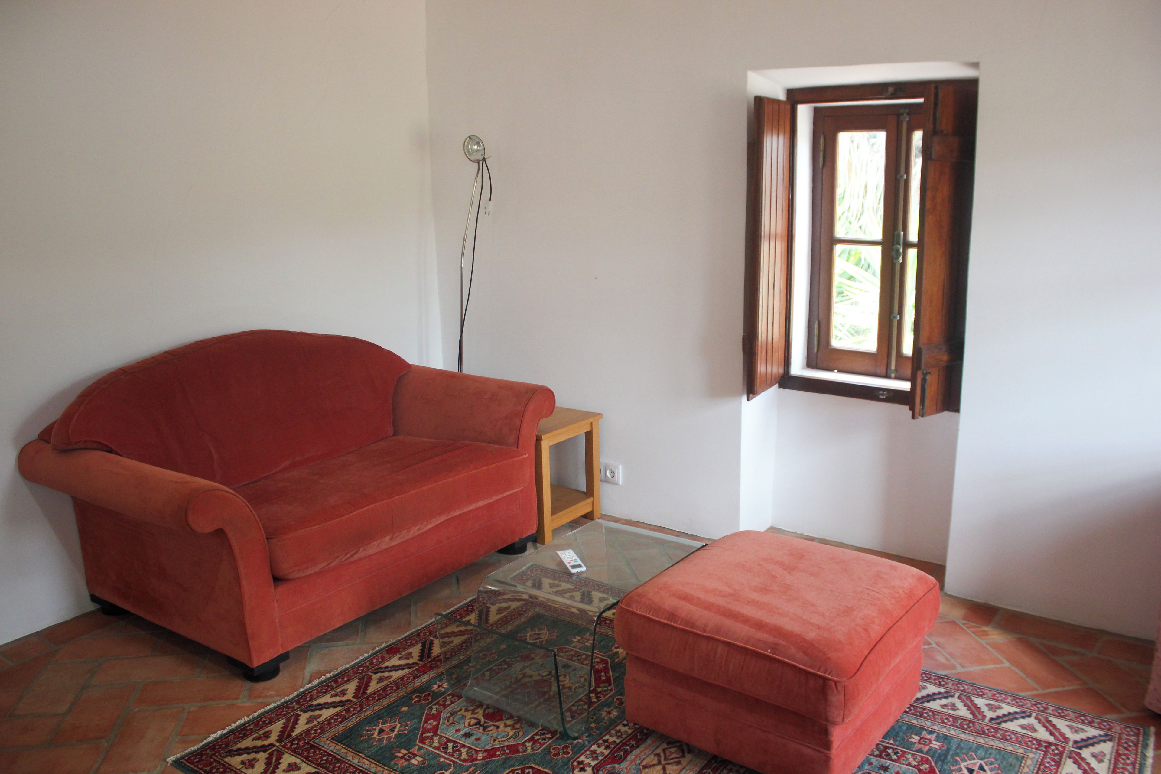 A room with a red sofa, a matching ottoman, a patterned rug, a small wooden table, and a window with a view of greenery.