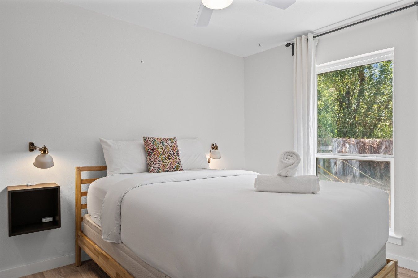 A bedroom with a bed, two bedside lamps, a window with curtains, and a view of trees.