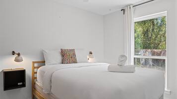 A bedroom with a bed, two bedside lamps, a window with curtains, and a view of trees.