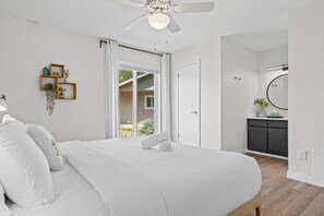 Un dormitorio con cama, ventilador de techo, ventana con cortinas, puerta, planta pequeña y espejo en la pared.