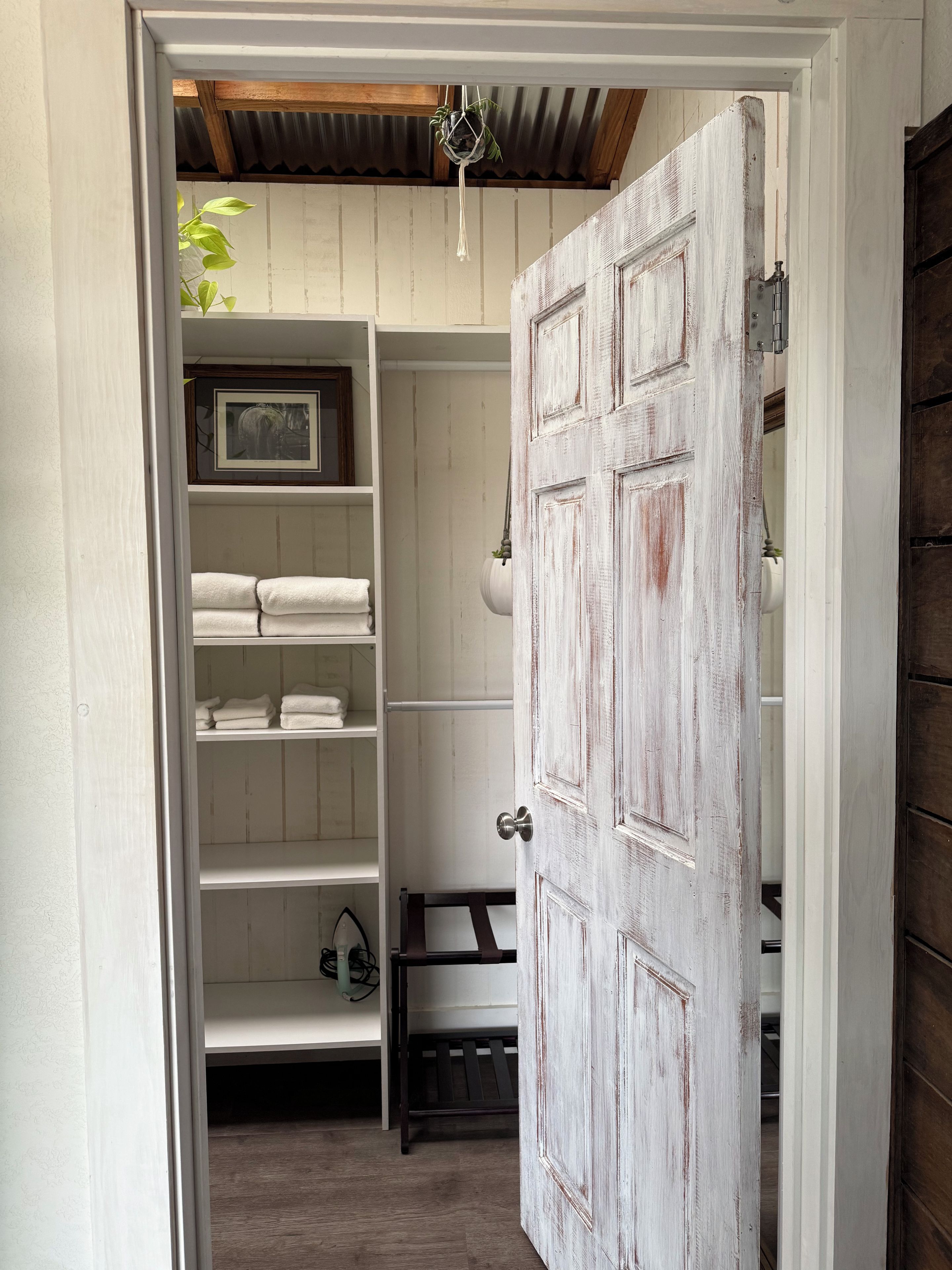 A hallway leading to a room with a rustic wooden door, a white shelving unit with folded towels, and a hanging plant.