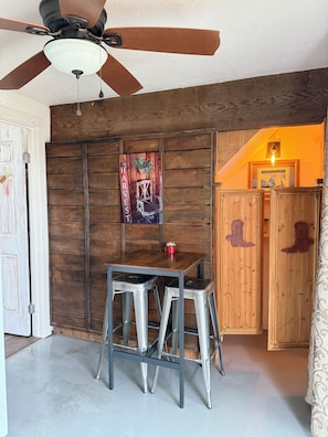 A room with a ceiling fan, a wooden wall with a 'HARVEST' sign, a table with stools, and a door leading to another room. - The Atrium- Templeton Ranch Retreat - Private Patio - Farm Animals- Wine Country (Templeton)