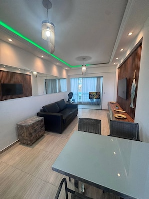 A modern living room with a sofa, a dining table, and a TV mounted on the wall. - Cumbuco Beach Place 02 – Piscina + 100 metros Mar (Caucaia)