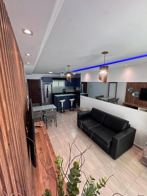Un salon moderne avec un canapé noir, un plancher en bois et un coin cuisine à l’arrière-plan.