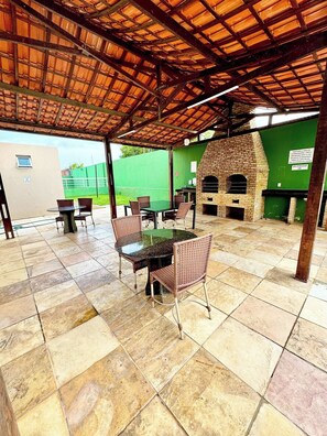 Eine Terrasse im Freien mit einem gemauerten Grill, Korbstühlen und einem Glastisch.