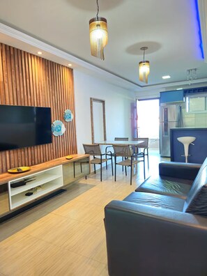 A modern living room with a flat-screen TV, wooden panel wall, dining area, and a kitchen with blue cabinets. - Cumbuco Beach Place 03 – Piscina + 100 metros Mar (Caucaia)