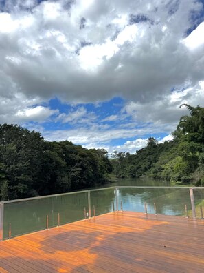 Una terrassa de fusta amb vista a un riu tranquil, amb un bosc dens a un costat i un cel serè a dalt.