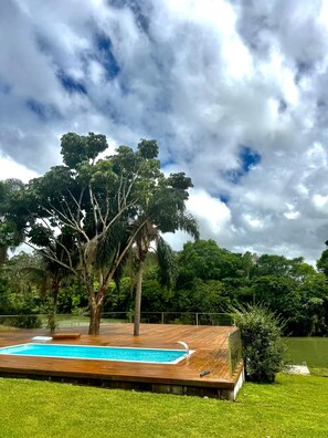 A swimming pool with a wooden deck, surrounded by lush greenery and a large tree.