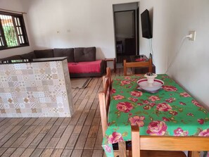 Zona de menjador amb una taula coberta per un mantell floral, cadires de fusta, un sofà amb un coixí vermell i una televisió penjada a la paret.