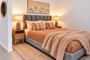 A modern bedroom with a grey upholstered bed, two bedside tables with lamps, a woven side table, a patterned rug, and a wall-mounted abstract painting. - Modern Luxury Miami Apartment with Pool & Gym (Miami)