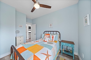 A bedroom with a bed, a nightstand, a dresser, a ceiling fan, and a door leading to another room. - Indian Beach | Decks, Views & Walk to Beach (Galveston)