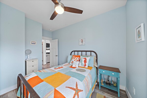 A bedroom with a bed, a nightstand, a dresser, a ceiling fan, and a door leading to another room. - Indian Beach | Decks, Views & Walk to Beach (Galveston)