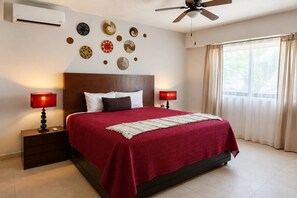 A hotel room with a bed, two bedside tables with lamps, a ceiling fan, a window with curtains, and wall decor. - 3BR Ground-Floor Condo with Pool & Terrace (Puerto Aventuras)